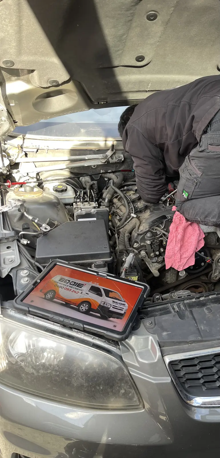 Mechanic working under the bonnet beside a tablet displaying Eddie The Mobile Mechanic